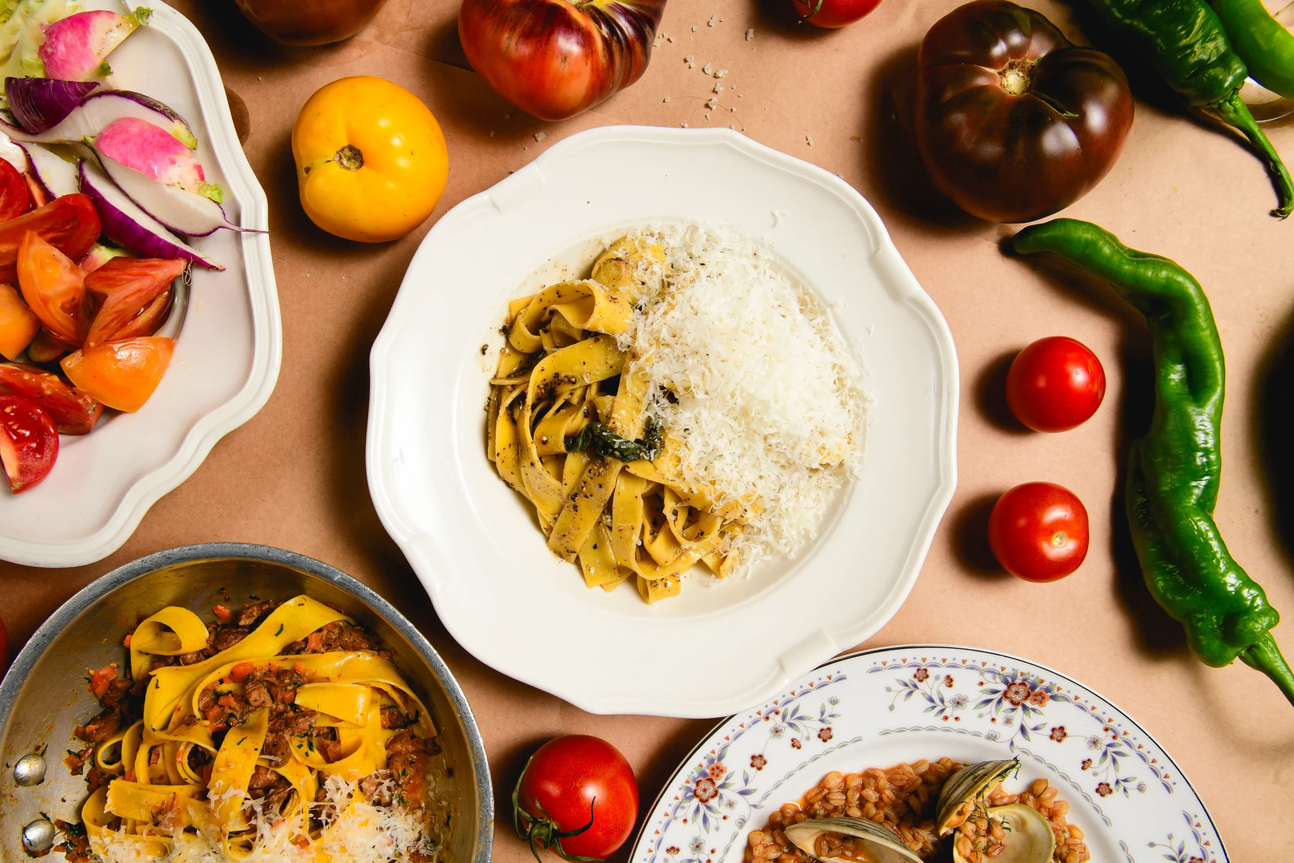 A table set with a plate of tagliatelle pasta topped with grated cheese, surrounded by fresh tomatoes, green peppers, radishes, and additional pasta and vegetable dishes.