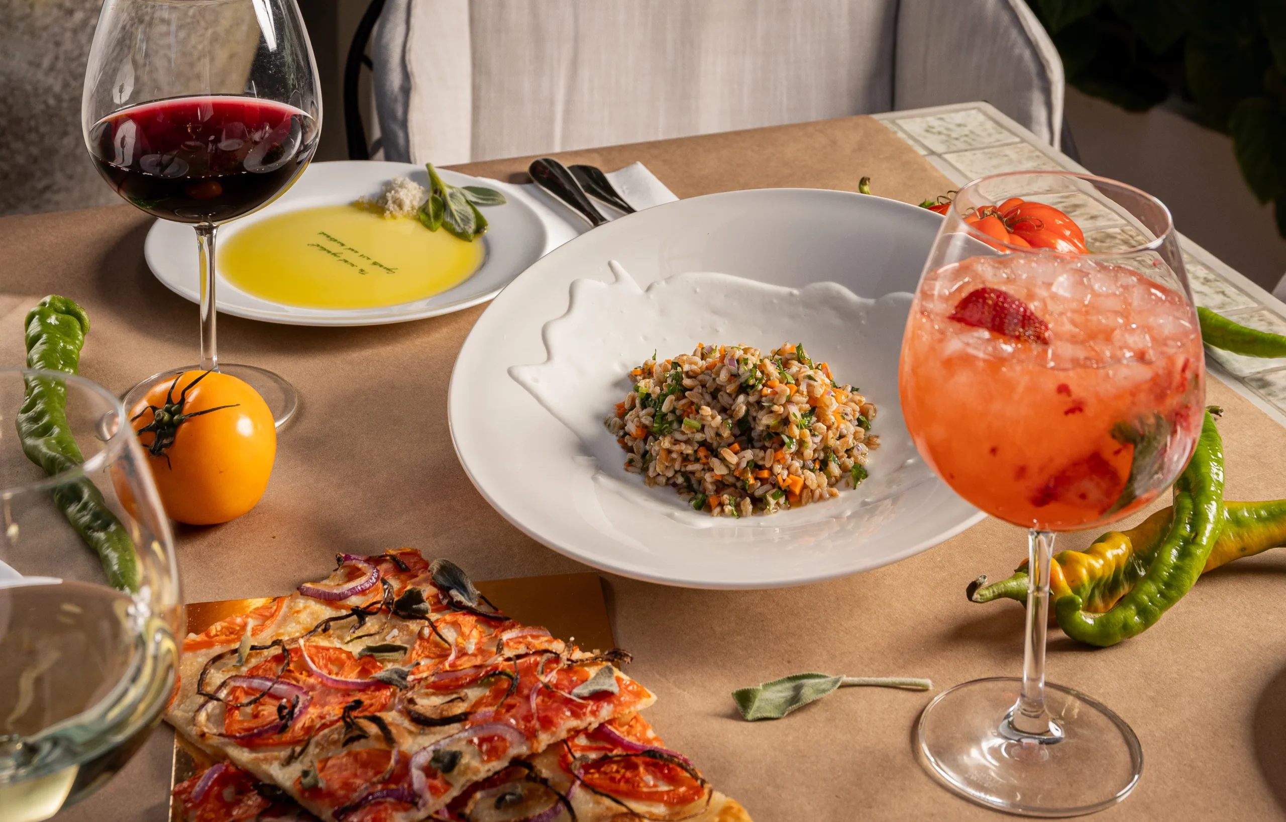A table set with a plate of mixed grain salad, a glass of red wine, a glass of strawberry cocktail, olive oil with herbs, fresh tomatoes and peppers, and a slice of tomato onion flatbread.
